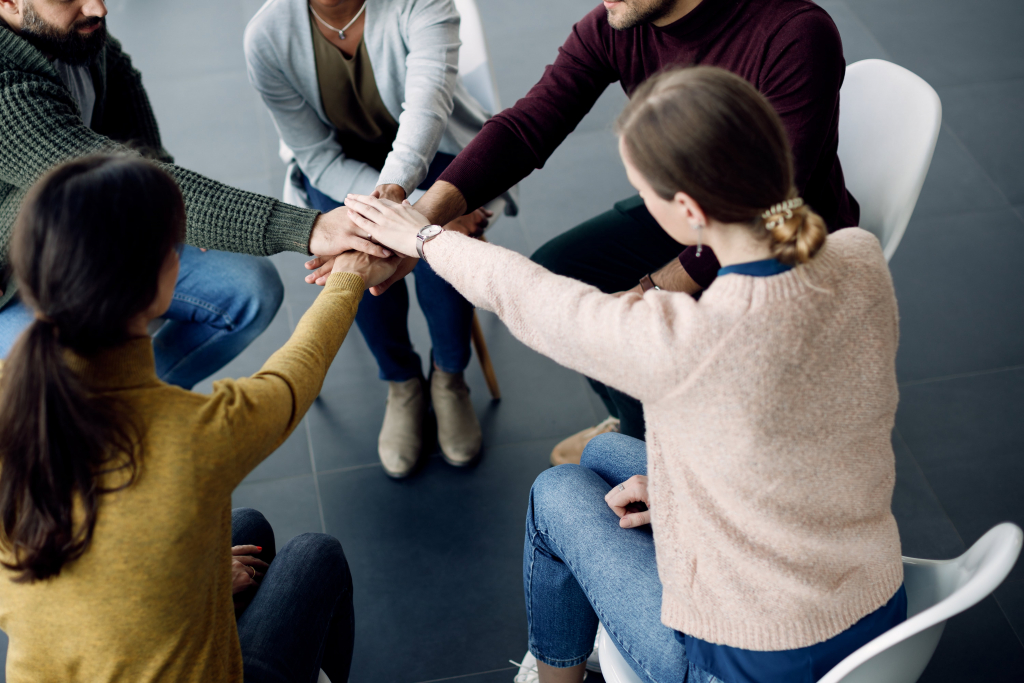 Fünf im Kreis sitzende Personen legen in der Mitte ihre Hände zusammen und suggerieren damit Teamarbeit oder Einigkeit.