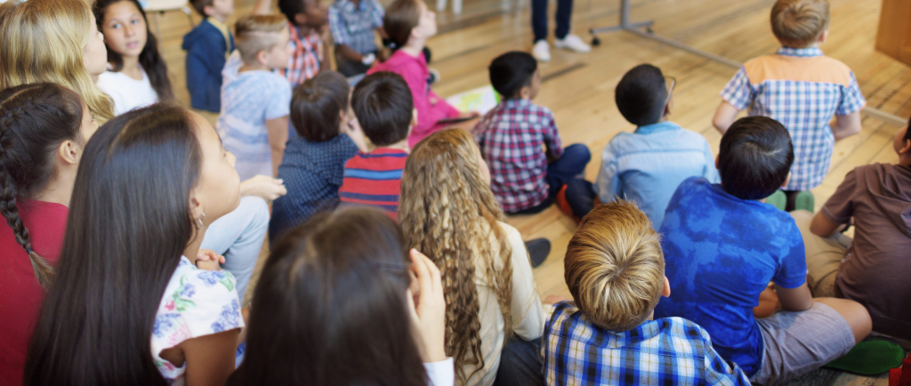 Eine Gruppe von Kindern sitzt in einem Klassenzimmer auf dem Boden und blickt einem Lehrer vorne im Raum zu.