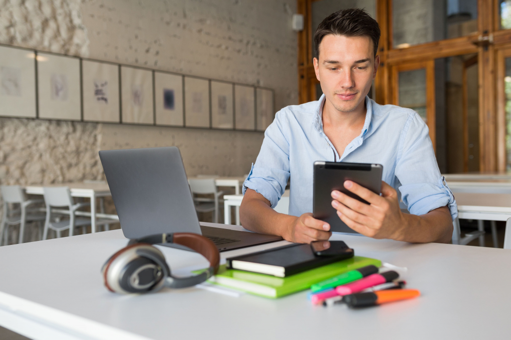 Eine Person sitzt mit Laptop, Tablet, Notizbüchern, Stiften und Kopfhörern an einem Tisch. Im Hintergrund sind braune Holzstühle und gerahmte Bilder zu sehen.