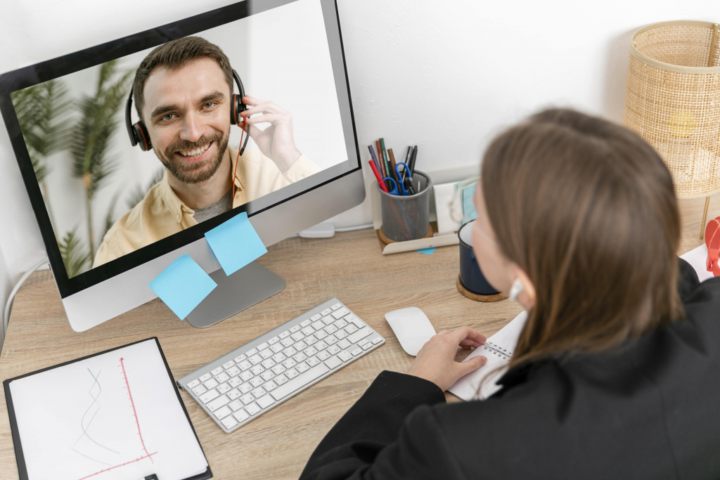 Eine Person mit Headset führt ein Videogespräch mit einer anderen Person, die an einem Schreibtisch mit Monitor, Tastatur und Schreibwaren sitzt.