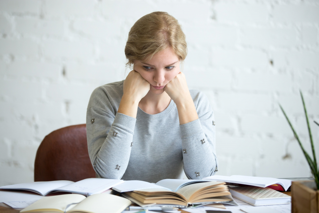 Frau sitzt an einem unordentlichen Schreibtisch, stützt ihr Kinn auf ihre Hände und blickt nachdenklich auf offene Bücher und Papiere.