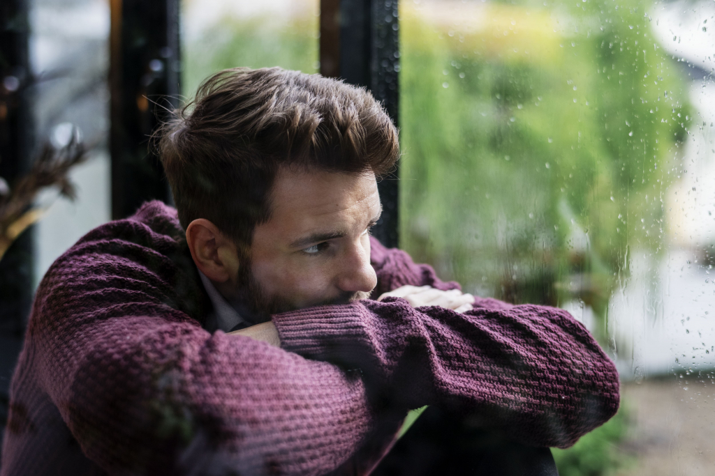 Ein Mann in einem kastanienbraunen Pullover lehnt an einem Fenster und blickt an einem regnerischen Tag nach draußen.