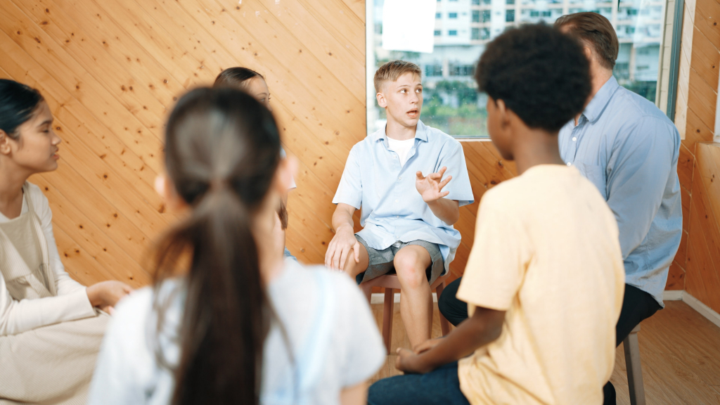 Eine Gruppe Teenagern, sitzt in einem Kreis in einem holzgetäfelten Raum.
