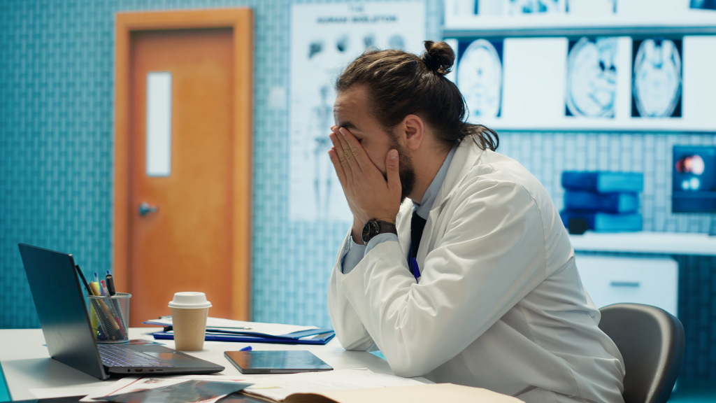 Ein Arzt im weißen Kittel sitzt an einem Schreibtisch und bedeckt sein Gesicht mit den Händen. Er wirkt gestresst. Auf dem Schreibtisch liegen Krankenakten und ein Laptop. Im Hintergrund sind Gehirnscans zu sehen.