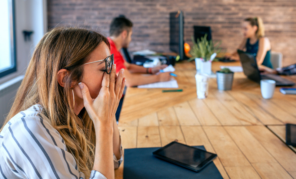 Eine Frau sitzt an einem Holztisch und bedeckt ihr Gesicht mit den Händen, während im Hintergrund einer Büroumgebung zwei weitere Personen an Laptops arbeiten.