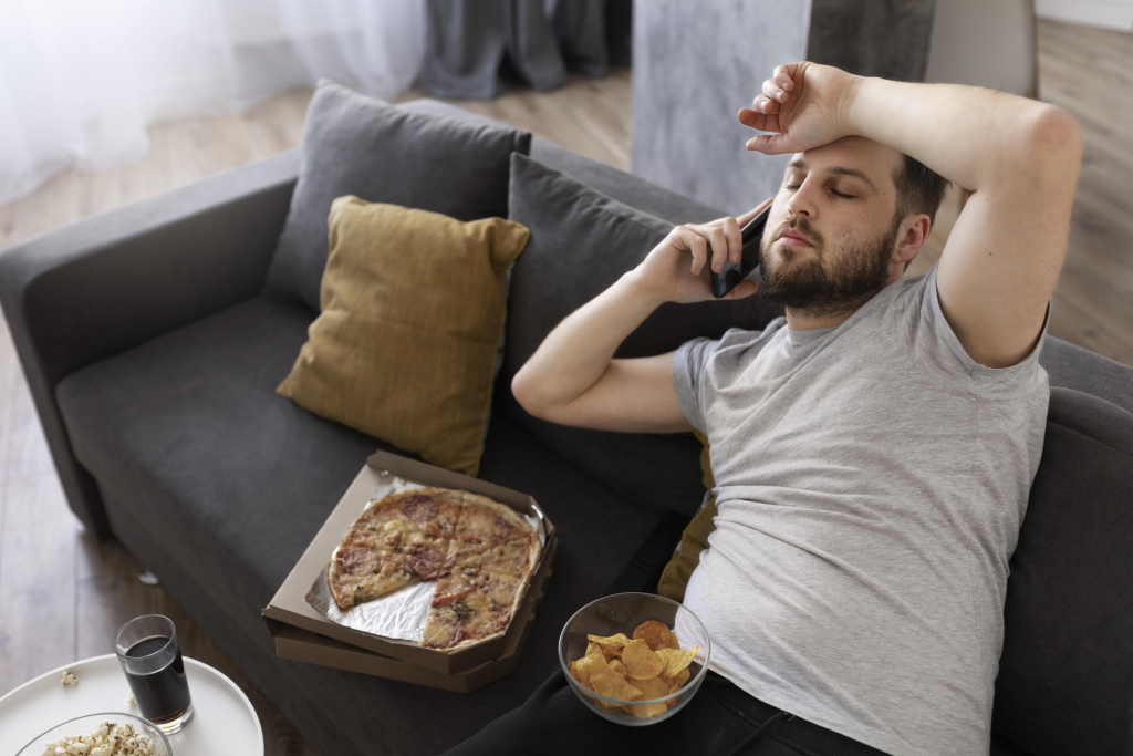 Ein Mann liegt auf einer Couch und hält sich ein Telefon ans Ohr. Auf dem Tisch daneben stehen eine Pizzaschachtel, Pommes und ein Glas Limonade.