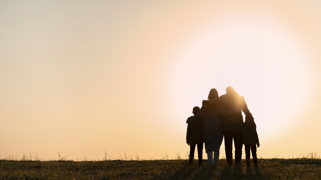 Eine vierköpfige Familie, von hinten gesehen, steht bei Sonnenuntergang im Freien zusammen, während die Sonne tief am Horizont steht.