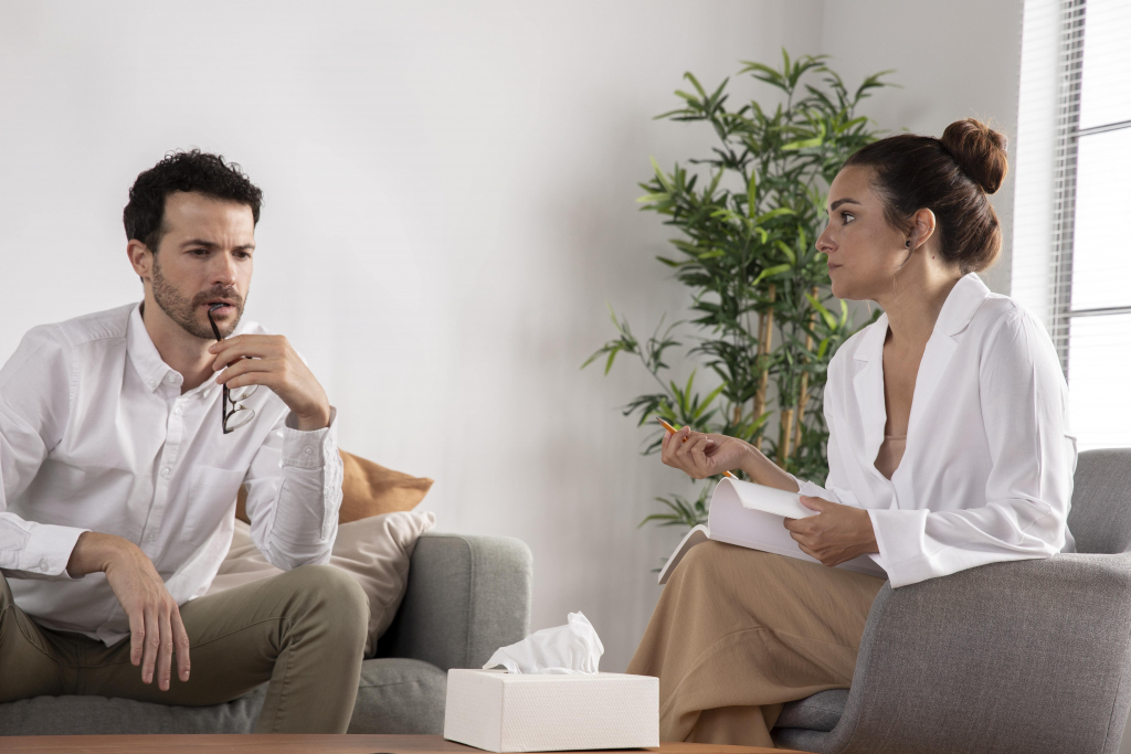 Ein Mann und eine Frau sitzen sich in einem hellen Raum gegenüber; die Frau hält ein Klemmbrett und scheint zuzuhören, was auf eine Therapie- oder Beratungssitzung hindeutet.