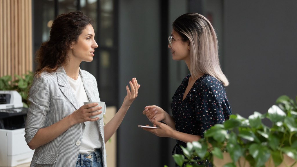 Zwei Frauen stehen in einem Haus und führen ein Gespräch; die eine hält einen Kaffeebecher, die andere ein Telefon. Im Hintergrund sind Grünpflanzen und Büromaschinen zu sehen.