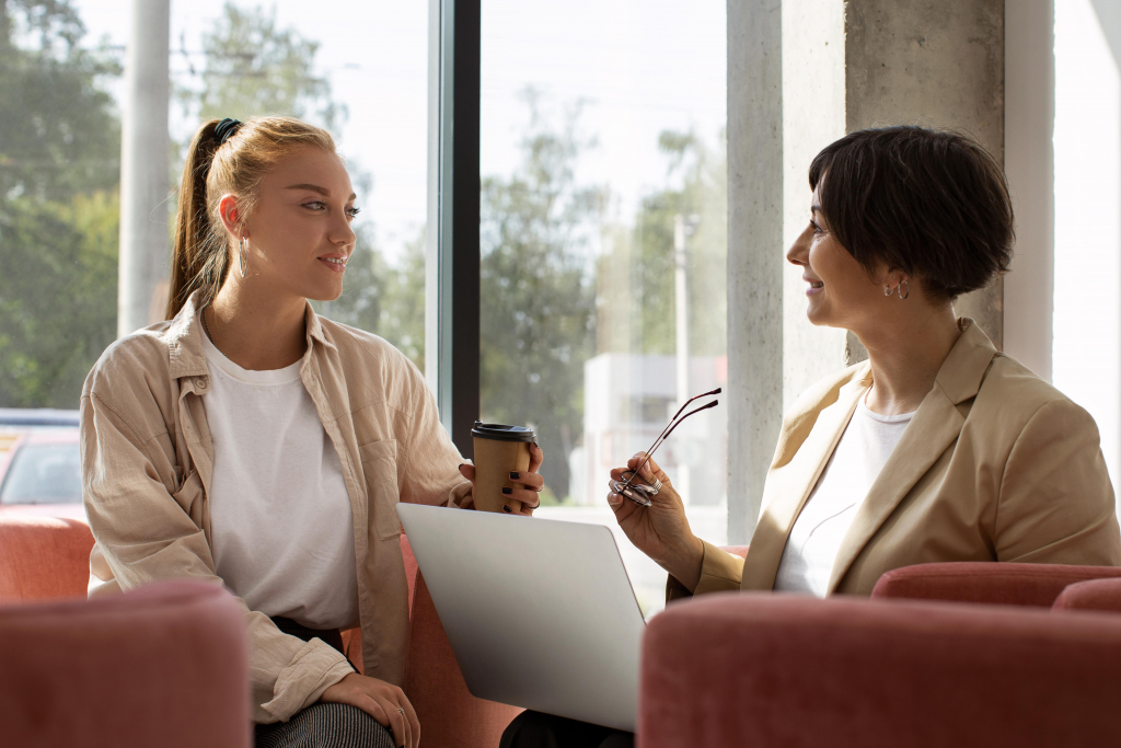 Zwei Frauen sitzen auf rosafarbenen Stühlen am Fenster, die eine mit einer Kaffeetasse, die andere mit einer Brille und einem Laptop in der Hand, und unterhalten sich.