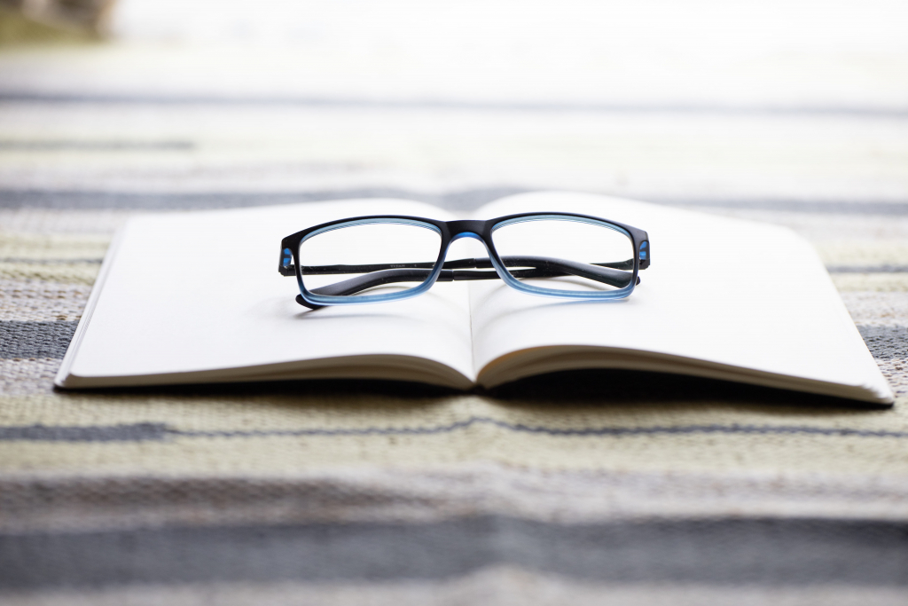 Eine schwarz umrandete Brille liegt auf einem aufgeschlagenen, leeren Notizbuch, das auf einer gestreiften Fläche liegt.