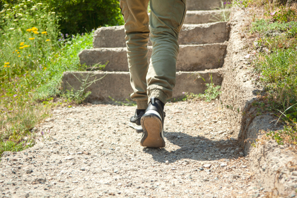 Eine Person in grünen Hosen und schwarzen Schuhen geht an einem sonnigen Tag eine Betontreppe hinauf, in deren Nähe sich Gras und gelbe Blumen befinden.