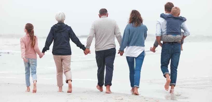 Sechs Personen unterschiedlichen Alters laufen barfuß über einen Strand, halten sich an den Händen und schauen von der Kamera weg. Ein Erwachsener trägt ein Kind. Der Himmel scheint bewölkt zu sein.