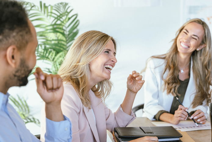 Drei Personen sitzen an einem Tisch und führen eine lebhafte Unterhaltung; zwei Frauen lächeln und lachen, während ein Mann aufmerksam zuhört. Im Hintergrund steht eine grüne Pflanze.